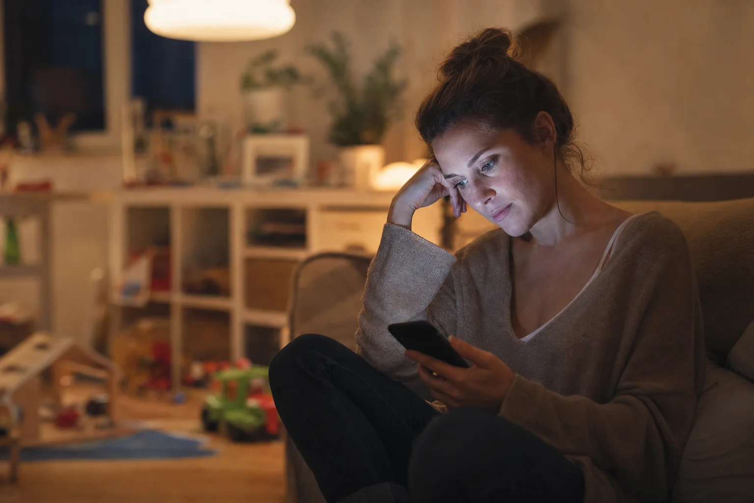Junge Mutter blickt abends nachdenklich auf ihr Smartphone und vergleicht sich mit anderen